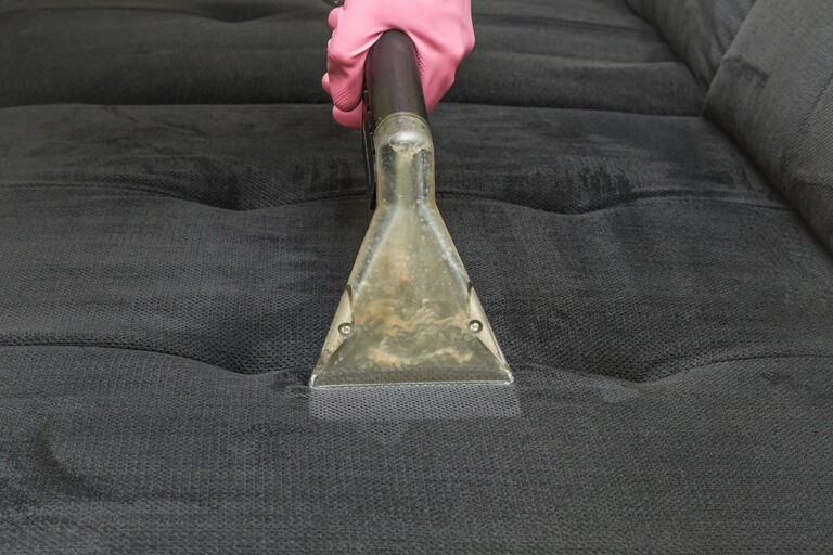 Ein aktiv Mitarbeiter reinigt ein anthrazitfarbenes Sofapolster im Rahmen einer Polsterreinigung mit einem Sauger und trägt dabei einen Gummihandschuh.